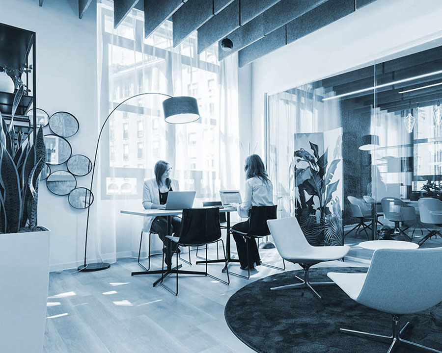 Two women sitting on a conference table in an office lounge decorated with plants and modern lighting.